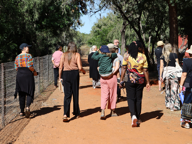 A photo of the back of a group of people, who are walking along a red dirt path in the bush with a fence line running along the side of them.